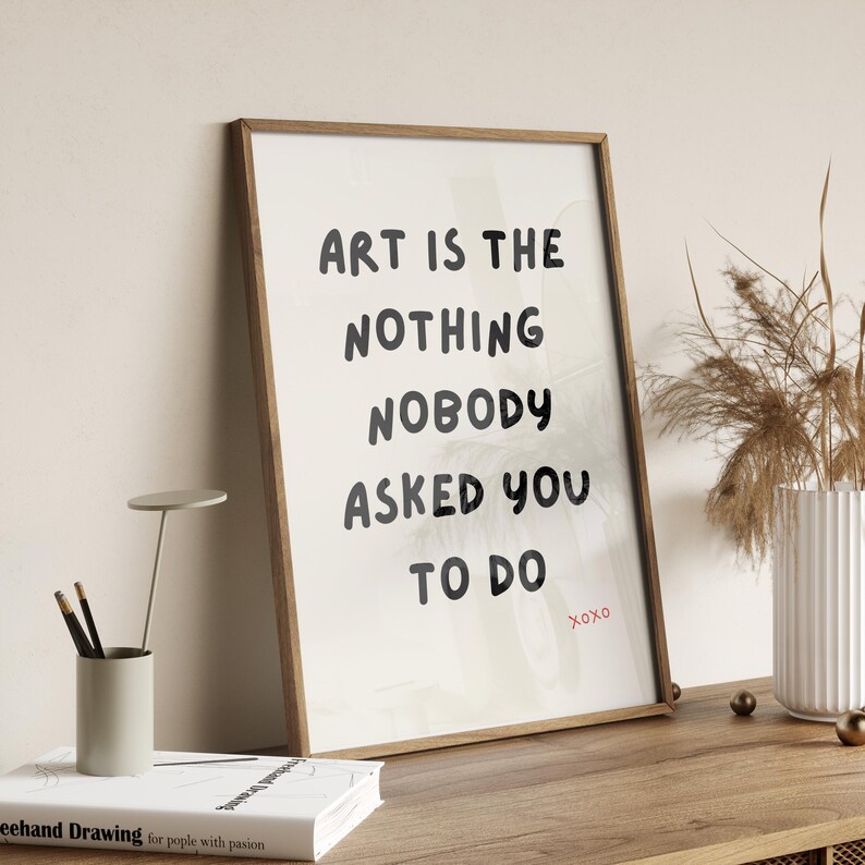 Op de afbeelding: Een ingelijst print met een witte achtergrond en zwarte tekst die "Art is the nothing nobody asked you to do" luidt, met het woord "Xoxo" in rood eronder.