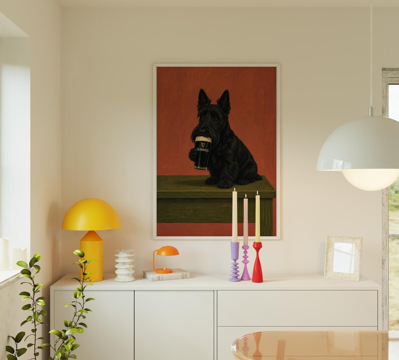 May include: A framed art print featuring a black Scottish Terrier dog holding a pint of Guinness. The dog is set against a red background, with a green table. The print is displayed above a white cabinet.