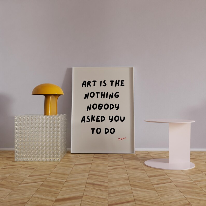Op de afbeelding: Een witte ingelijste print met de tekst "Art is the nothing nobody asked you to do xoxo" op een beige achtergrond. De print staat op een houten vloer met een gele lamp en een witte tafel.