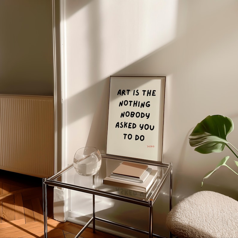 Op de afbeelding: Een ingelijst print met de tekst "Art is the nothing nobody asked you to do" op een witte achtergrond. De print staat op een glazen tafel met een glazen kom en een stapel boeken.
