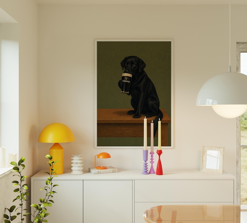 Pu&ograve; includere: Una stampa incorniciata con un Labrador Retriever nero che tiene in mano una pinta di Guinness. Il cane &egrave; seduto su una superficie di legno su uno sfondo verde scuro. La stampa &egrave; esposta sopra un mobile bianco con candele e una lampada gialla.