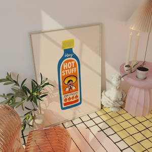Puede incluir: Un cuadro enmarcado con una ilustración de una botella de salsa picante. La botella es azul con una tapa amarilla y las palabras "Hey HOT STUFF". Debajo, una imagen de dibujos animados de un hombre con sombrero. La impresión se muestra en una habitación.
