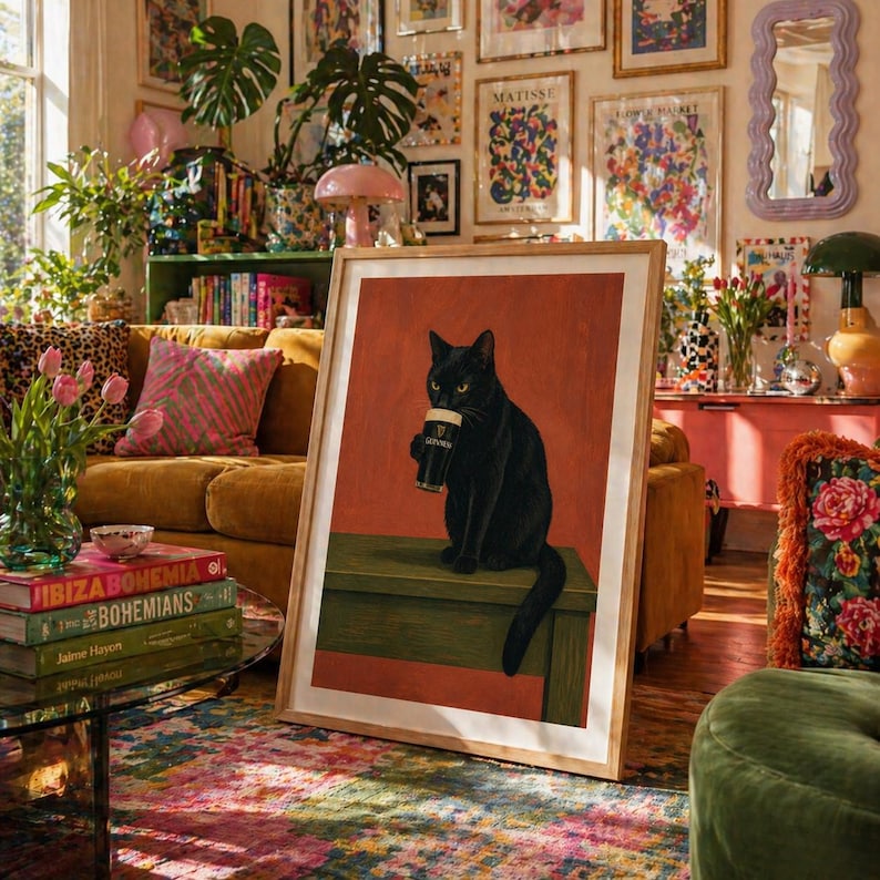 Puede incluir: Un cuadro enmarcado con un gato negro sosteniendo una pinta de Guinness. El gato est&aacute; sentado sobre una superficie verde contra un fondo rojo-naranja. La impresi&oacute;n est&aacute; en un marco de madera, creando un ambiente c&aacute;lido.