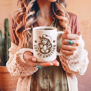 Puede incluir: Una taza de cer&aacute;mica blanca con un gr&aacute;fico marr&oacute;n de una persona con sombrero de vaquero sosteniendo una taza peque&ntilde;a, rodeado por el texto "CUP OF AMBITION" y dos estrellas. La taza tiene un asa resistente y contiene un l&iacute;quido oscuro.