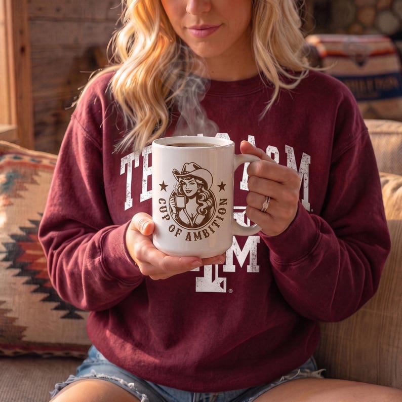 Puede incluir: Una taza de cer&aacute;mica blanca con un gr&aacute;fico marr&oacute;n de una mujer con sombrero de vaquero que sostiene una taza peque&ntilde;a, rodeada por el texto "CUP OF AMBITION" con dos estrellas. La taza se sostiene con ambas manos y sale vapor de ella.