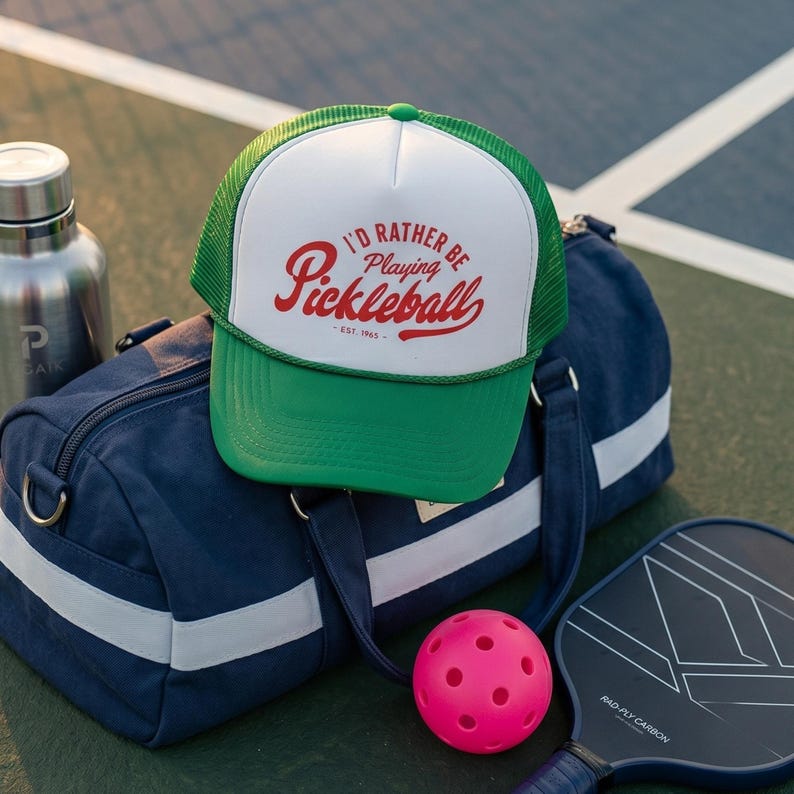 Puede incluir: Gorra de camionero verde y blanca con el texto "I'd Rather Be Playing Pickleball - Est. 1965" en escritura roja. La gorra est&aacute; colocada sobre una bolsa de lona azul marino con rayas blancas, una pelota de pickleball rosa y una pala de pickleball.