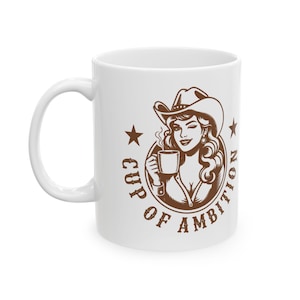 Puede incluir: Una taza de cer&aacute;mica blanca con una ilustraci&oacute;n gr&aacute;fica marr&oacute;n de una persona con sombrero de vaquero que sostiene una taza humeante. El dise&ntilde;o est&aacute; encerrado en un marco circular con dos estrellas y el texto "CUP OF AMBITION" en letra negrita.
