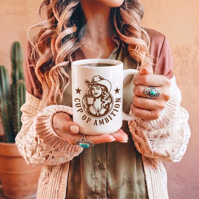 Puede incluir: Una taza de cer&aacute;mica blanca con un gr&aacute;fico marr&oacute;n de una persona con sombrero de vaquero sosteniendo una taza peque&ntilde;a, rodeado por el texto "CUP OF AMBITION" y dos estrellas. La taza tiene un asa resistente y contiene un l&iacute;quido oscuro.