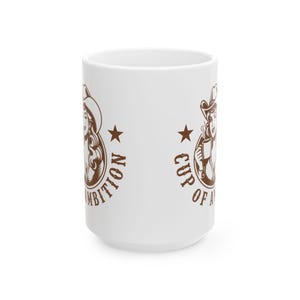 Puede incluir: Una taza de cer&aacute;mica blanca con un gr&aacute;fico marr&oacute;n de una persona con sombrero de vaquero que sostiene una taza humeante. El dise&ntilde;o incluye una estrella y el texto "CUP OF AMBITION" impreso en un dise&ntilde;o circular alrededor de la ilustraci&oacute;n.