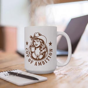 Puede incluir: Una taza de cer&aacute;mica blanca con un gr&aacute;fico marr&oacute;n de una persona con sombrero de vaquero que sostiene una taza humeante. El texto "CUP OF AMBITION" est&aacute; impreso en un arco circular alrededor de la ilustraci&oacute;n, flanqueado por dos estrellas.
