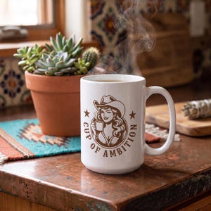 Puede incluir: Una taza de cer&aacute;mica blanca con un gr&aacute;fico marr&oacute;n de una persona con sombrero de vaquero sosteniendo una taza peque&ntilde;a, rodeado por el texto "CUP OF AMBITION" y dos estrellas. El vapor sale de la taza situada sobre una superficie de color cobre.