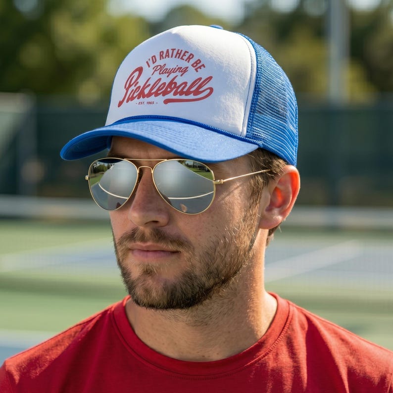 Puede incluir: Gorra de camionero azul y blanca con el texto "I'D RATHER BE PLAYING Pickleball" en escritura roja. La gorra tiene una visera azul y una parte trasera de malla. La persona de la imagen lleva gafas de sol aviador doradas y una camiseta roja.