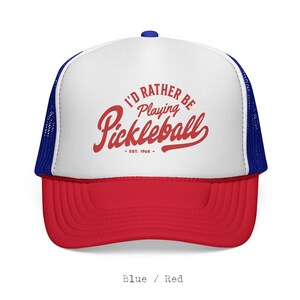 Puede incluir: Gorra de camionero roja, blanca y azul con el texto "I'D RATHER BE PLAYING PICKLEBALL - EST. 1965" en escritura roja. La gorra tiene una visera roja, un panel frontal blanco y una parte trasera de malla azul. Un bot&oacute;n azul est&aacute; en la parte superior.