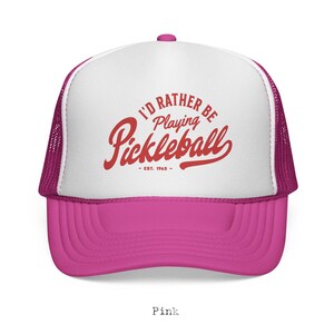 Puede incluir: Gorra de camionero rosa y blanca con el texto "I'd Rather Be Playing Pickleball" en escritura roja. La gorra tiene una visera rosa y una parte trasera de malla. El texto "EST. 1965" est&aacute; debajo.