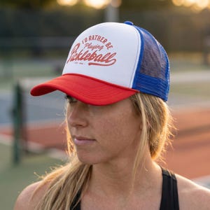 Puede incluir: Gorra de camionero roja, blanca y azul con el texto "I'D RATHER BE playing Pickleball" en escritura roja. La gorra tiene una visera roja, un panel frontal blanco y una parte trasera de malla azul. El texto "EST. 1969" est&aacute; debajo.