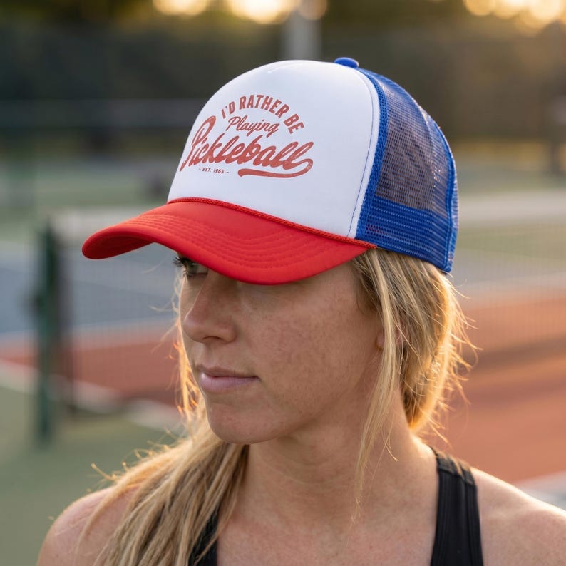 Puede incluir: Gorra de camionero roja, blanca y azul con el texto "I'D RATHER BE playing Pickleball" en escritura roja. La gorra tiene una visera roja, un panel frontal blanco y una parte trasera de malla azul. El texto "EST. 1969" est&aacute; debajo.
