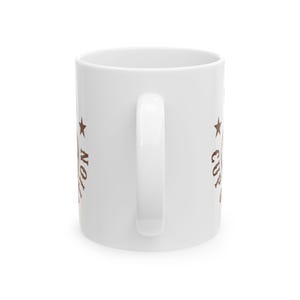 Puede incluir: Una taza de cer&aacute;mica blanca con un dise&ntilde;o gr&aacute;fico marr&oacute;n en ambos lados. El dise&ntilde;o incluye un s&iacute;mbolo de estrella y el texto "CUP OF AMBITION" en una fuente de estilo vintage desgastada. La taza tiene un acabado liso y brillante.