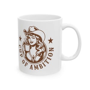 Puede incluir: Una taza de cer&aacute;mica blanca con un gr&aacute;fico marr&oacute;n de una persona con sombrero de vaquero que sostiene una taza humeante. El dise&ntilde;o est&aacute; encerrado en un marco circular con una estrella a cada lado y el texto "CUP OF AMBITION" en letra negrita.