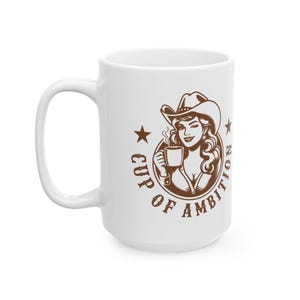 Puede incluir: Una taza de cer&aacute;mica blanca con un gr&aacute;fico marr&oacute;n de una persona con sombrero de vaquero que sostiene una taza humeante. El dise&ntilde;o circular incluye dos estrellas y el texto "CUP OF AMBITION" impreso en una fuente negrita y desgastada.