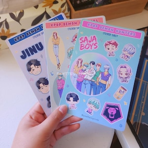 May include: Three KPOP Demon Hunters sticker sheets. One sheet features the name "JINU" with cartoon faces and musical notes. Another sheet has a drawing of a person. The third sheet has the text "SAJA BOYS" with cartoon illustrations of people and a lion logo.