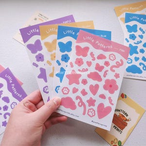Peut inclure: Plusieurs planches d'autocollants "Little Patterns" sont présentées. Les autocollants, tenus par une main, affichent des papillons, des fleurs, des étoiles, des nuages, des empreintes de pattes et des cœurs en violet, jaune, bleu et rose.