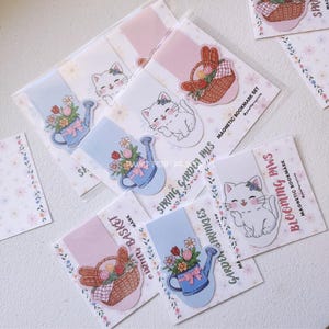 Può includere: Un set di segnalibri magnetici a tema giardino primaverile. I segnalibri presentano illustrazioni di un gatto bianco, annaffiatoi e cesti pieni di fiori. Il testo "Spring Garden Pals" e "Blooming Paws" è visibile.