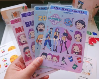Op de afbeelding: Een hand houdt verschillende KPOP-stickerbladen vast. De bladen bevatten kleurrijke illustraties van personages in anime-stijl en de tekst "KPOP DEMON HUNTERS" en verschillende namen. De stickers zitten in doorzichtige plastic hoezen.