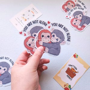May include: A hand holds several stickers featuring cartoon monkeys hugging. The stickers have the text "YOU ARE NOT ALONE" and small heart accents. Additional stickers and a business card with a cat illustration are also visible.