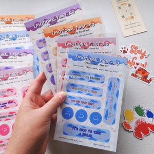 May include: A collection of "Emotional First-Aid" sticker sheets in various colors, including purple, orange, pink, and blue. Each sheet features uplifting phrases and designs, such as "It's okay to take a break." The stickers are packaged in clear plastic.