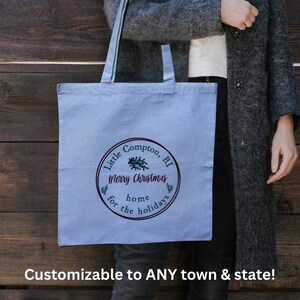May include: A light blue tote bag with a circular design that reads "Little Compton, RI Merry Christmas for the home holidays." The bag has long straps and is held by a person. The text "Customizable to ANY town & state!" is at the bottom.
