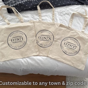 Puede incluir: Tres bolsas de mano beige con diseños circulares negros con nombres de ciudades y códigos postales de Rhode Island. El texto "Customizable to any town & zip code!" está en la parte inferior.