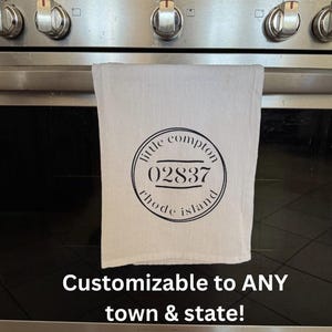May include: A white kitchen towel with a black circular design featuring the text "little compton 02837 rhode island." Below the towel is the text "Customizable to ANY town & state!" The towel is hanging on a stainless steel oven.