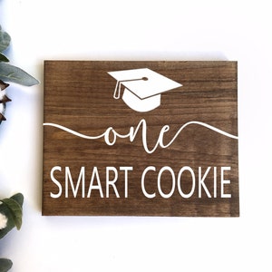 May include: A brown wooden sign with white text that reads "one smart cookie". A white graduation cap is above the text.