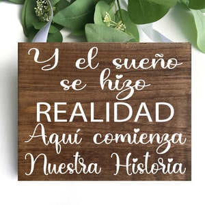 Könnte beinhalten: Ein quadratisches, dunkelbraunes Holzschild mit weißer Schrift. Der spanische Text lautet "Y el sueño se hizo REALIDAD Aquí comienza Nuestra Historia". Das Schild wird von einem weißen Band gehalten. Grünes Laub ist im Hintergrund zu sehen.