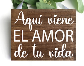 May include: A rectangular, wooden sign with the Spanish text "Aquí viene el amor de tu vida" in white script. The sign is a dark brown color and has a white ribbon for hanging. Greenery and white ribbon are in the background.