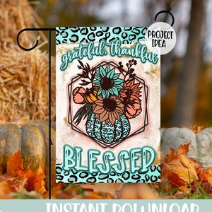 May include: A rectangular garden flag featuring a teal leopard print border, a central geometric frame containing a bouquet of sunflowers and pumpkins, and the text "grateful thankful" at the top and "BLESSED" in large teal letters at the bottom.