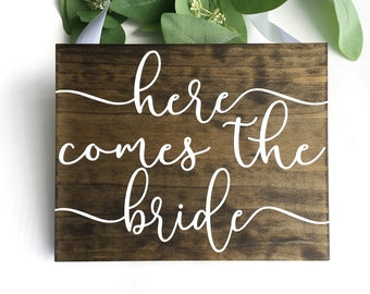 May include: A wooden sign with the text "here comes the bride" in white cursive lettering on a brown background.