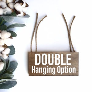 May include: A wooden sign with the text "DOUBLE Hanging Option" in white lettering. The sign has two pieces of twine attached to the top for hanging.
