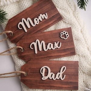 Puede incluir: Tres etiquetas de regalo de madera con las palabras "Mom", "Max" y "Dad" en fuente cursiva. La etiqueta "Max" incluye una huella de pata. Cada etiqueta es marrón con una cuerda de yute para colgar. Las etiquetas descansan sobre una superficie de punto blanca.