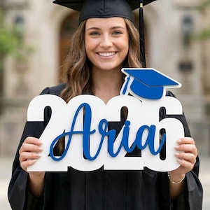 以下が含まれることがあります： 黒いガウンと帽子を身に着けた卒業生が、青い文字で「Aria 2026」と書かれた白いサインと青い卒業キャップを持っています。背景にはぼやけた建物があります。