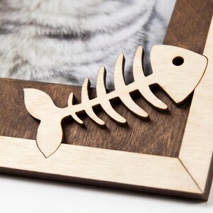 May include: A wooden picture frame featuring a two-tone design with a dark brown inner border and a light beige outer border. A light-colored wooden cutout of a fish skeleton is attached to the corner of the frame.