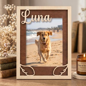 May include: A rectangular wooden picture frame with a light wood outer border and a dark wood inner frame. The top features the name Luna in script, and the bottom corners have carved angel wing designs. It holds a photo of a golden dog.