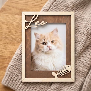 May include: A rectangular wooden picture frame with a dark brown inner border and a light wood outer frame. The name Leo is cut out in light wood at the top left, and a fish skeleton shape is attached at the bottom right corner.