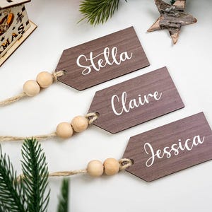 Puede incluir: Tres etiquetas de madera marrón con los nombres "Stella", "Claire" y "Jessica" en escritura blanca. Cada etiqueta está unida a una cuerda con cuentas de madera. Ideales para la decoración navideña u otras festividades.