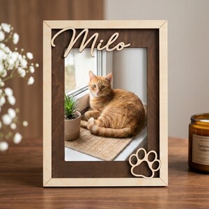 May include: A rectangular wooden picture frame with a light wood outer border and a dark wood inner frame. The top features the name Milo in script, and the bottom right corner has a cutout paw print. The frame holds a photo of an orange cat.