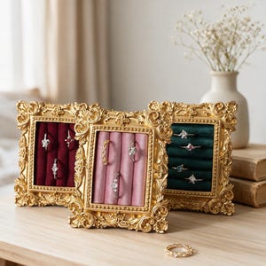 May include: Three rectangular jewelry display stands with ornate gold frames. Each stand features a velvet-lined interior with padded slots for rings. The velvet colors are deep red, soft pink, and dark green, holding various silver-toned rings.