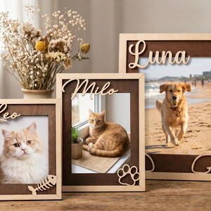 May include: Three rectangular wooden picture frames with dark brown inner borders and light wood outer edges. The frames feature laser-cut names Leo, Milo, and Luna, with decorative accents including a fish skeleton, paw print, and angel wings.