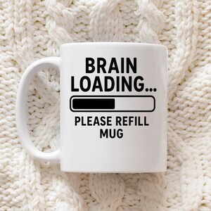 Puede incluir: Taza de cerámica blanca con el texto "BRAIN LOADING..." encima de una barra de carga y "PLEASE REFILL MUG" debajo. La taza tiene un asa blanca y está sobre un fondo texturizado blanquecino.