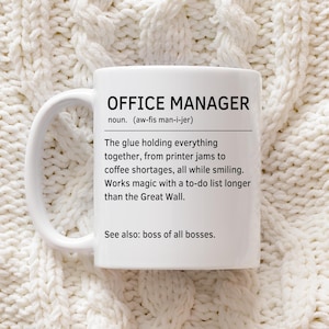 May include: White ceramic mug with the text "OFFICE MANAGER" in black. The mug includes a definition and humorous description of an office manager. The mug is on a white knitted surface.