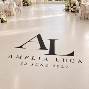 May include: A white dance floor decal featuring the large black monogram letters AL, with the names AMELIA and LUCA printed below in a serif font, followed by the date 12 JUNE 2027 in black text centered on the smooth floor surface.